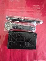 Black pen set in plastic packaging, unbranded wrist watch with black strap and large white numerals, and black faux crocodile texture credit/business card case.