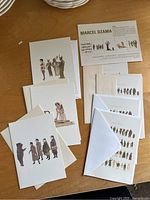 Photos showing a collection of illustrated art cards and white envelopes on a wooden surface, featuring different stylized human and animal characters by Marcel Dzama.