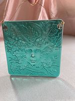 Front view of teal green square art glass light catcher showing embossed face and leaf design, wire hanger with metal charms attached at corners