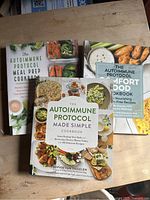 Three autoimmune protocol cookbooks stacked on a wooden surface, showing colorful covers with images of healthy dishes and ingredients.