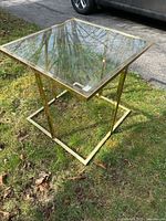 Side view of brass frame glass side table on grass with a car and driveway in background.