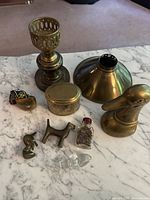 Lot arranged on marble surface showing brass candlestick, brass shade, brass figurines, small brass box, ring, glass bottle with cork, and small clear glass bird figurine.