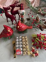 Full view of Christmas decor lot items arranged on a table including two velvet reindeer, wooden nutcracker, two red cardinal bird ornaments, pine cone and berry garland, and boxes of gold and silver mini ball ornaments.