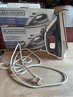 Front-angle photo of the Black+Decker Light 'N Easy compact steam iron placed upright on a wooden surface with the original product box behind it. The iron has a white and black body, visible controls, and attached white power cord.