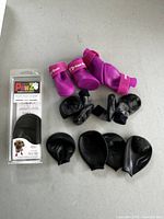 Overview of dog boots including unopened Pawz packaging, three pairs of used black boots, and two pairs of pink boots with Velcro straps.