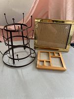 Metal mug holder rack next to brass-colored and wooden picture frames on neutral surface with pink fabric backdrop.