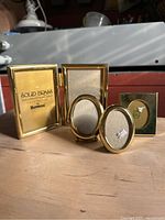 Five vintage solid brass small picture frames arranged on wooden surface with background showing some red painted and metal surfaces