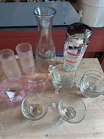 Photo showing a glass cocktail shaker with metal lid missing cap, a clear glass carafe, tall ribbed glasses, pink faceted glasses, small clear bowls, and punch cups.