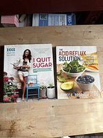 Front covers of two cookbooks, "I Quit Sugar" and "The Acid Reflux Solution," displayed on a wooden table