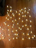 Full view of the curtain string lights laid out on a wooden floor showing multiple lit globe-shaped light covers connected by white wiring.