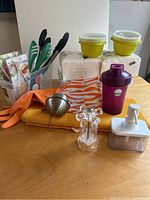 Photo showing assorted kitchen items on a wooden table: silicone gloves, tongs, tea infuser, green containers, folded towels, shaker bottle, spice grinder, and dispenser container.