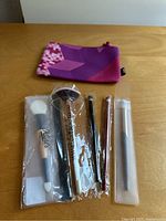 Photo showing set of six makeup brushes in plastic sleeves in various sizes and shapes, alongside purple pink patterned makeup pouch on wooden surface.
