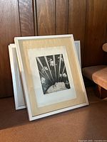 Framed pencil art in white IKEA frame leaning against wooden wall, beige matting surrounding pencil drawing.