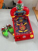 Overview of the mini pinball game with attached gumball dispenser, showing the red body and two green character slippers placed beside it.