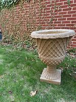 Full view of the fibreglass faux stone urn planter on grass with brick wall background.