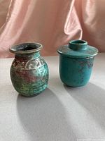 Two distinct Raku pottery pieces on a beige surface with a pink fabric background, showing a small rounded vase and a lidded jar from the front.