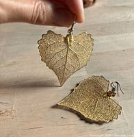 Photo of hand holding one of two gold dipped leaf earrings, showing delicate leaf veins and natural edges with hook attachment.