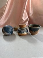 Four signed vintage studio pottery items shown including two small pots, a bowl, and a decorative ball, all in blue and earthy glazes on cloth background.