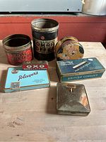The photo shows six vintage advertising tins and boxes on a wooden surface. Various brands including Okaro Coffee, Imperial Mixture, Oxo Cubes, Player's cigarettes, British Consols, and a round tin with a cartoon child face are visible.