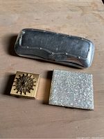 Top view of vintage glasses case, pill box with ornate sunflower design, and a compact with pearlescent finish.