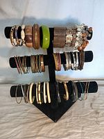 Full view of over 45 assorted bracelets on a black multi-tier stand showing variation in sizes, colors and materials including wood, metal, resin, and enamel.