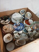 Box containing a variety of miniature tea set pieces including cups, saucers, plates, and pots with various colors and styles.