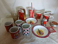 Photo showing assorted Coca-Cola mugs, plates, glass, plastic cup, vending machine replica, tape dispenser, and tape roll on white background.