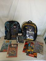 Photo showing two Batman backpacks, one blue and one black with yellow accent, Batman bobblehead in packaging, and seven Batman comics laid out.