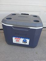 Front view of the blue and gray Coleman cooler with four cup holders on the lid. The cooler is solid blue with the Coleman logo and label visible on the front side.