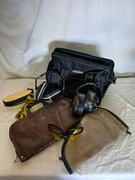 Photo showing black sportsman's bag, two pairs of shooting glasses with yellow lenses, two brown leather holsters, and one pair of earmuffs on a light fabric background.