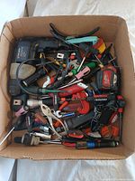 Top view of cardboard box filled with various hand tools including drills, screwdrivers, pliers, utility knives, sockets, and measuring tools.