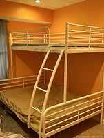 Photo showing white metal bunk bed against an orange wall with metal guardrails on top and bottom bunks and a slanted ladder for access to the top bunk, mattresses included.