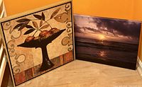 Framed artwork showing a pedestal bowl with five apples and leaf designs, beside a canvas print of a sunset over the ocean.