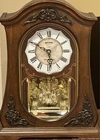 Front view of Buxton pendulum wall clock with ornate dark wood case, white dial with Roman numerals, and visible pendulum through glass window