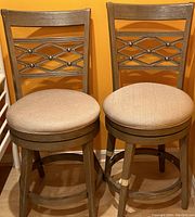Image showing two wooden bar chairs with beige cushioned seats and decorative metal lattice designs on each backrest. One chair has a broken leg wrapped with tape for stabilization.