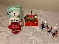 Photo showing four Christmas ornaments: Boyds plush bear in stocking, carousel exotic cat, and two Flintstones plastic bell ornaments.