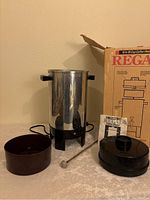 Stainless steel coffee maker body, black plastic lid, brown container, metal stir stick, instruction manual, and box shown on table.