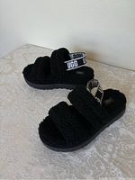 Side view of black fuzzy Ugg sandal showing plush straps and branded heel strap