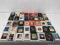 Top-down view of 38 assorted vintage 8 track cassette tapes laid out on a flat surface in rows. Tapes vary in color and condition.