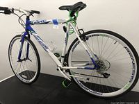 Side view of the Genesis GS700 hybrid bicycle showing the full white and blue frame, with visible Genesis branding on the frame and rims, green pedals, and cable lock.