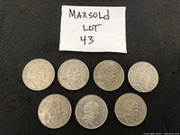 Seven Mexican Cinco Pesos coins arranged on a dark surface under paper with "MAXSOLD LOT 43" visible.