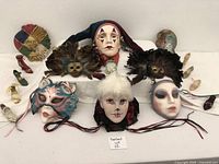 Collection of five masks and faces arranged on white cloth, showing variations in color and style including feathers and ribbons.