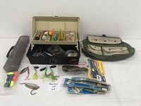 Full view of open beige fishing tackle box filled with assorted fishing lures, bobbers, hooks and other fishing accessories, along with green pouch and additional packaged fishing lures and accessories on a white surface.