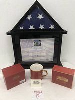 Photo of flag in triangular fold inside black display case with certificate below and presidential cup with original box and booklet in front.