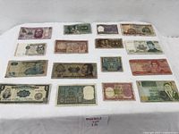 Top-down photo showing 16 mixed foreign used paper currencies laid out on a white fabric surface, light wear and fold marks visible on many notes.