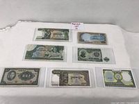 Overview of 7 assorted paper currency notes from different countries placed on a white cloth, each in a protective sleeve.