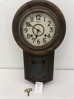 Front view of a vintage Hudson clock with round face, white dial with black numbers and hands, wooden frame, and winding key placed next to it.