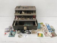 Front view of open tackle box displaying contents including hooks, sinkers, reels, fishing lines, and other accessories arranged in compartments and outside the box.