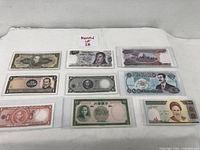 Eight uncirculated world banknotes laid flat, individually sleeved, showing front faces with portraits and denominations from various countries.