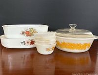 Four white glass custard cups with yellow Wheat pattern stacked, alongside three vintage Fire King bakeware pieces: one round casserole with glass lid and yellow daisy pattern, and two rectangular baking dishes with different floral and fruit patterns.